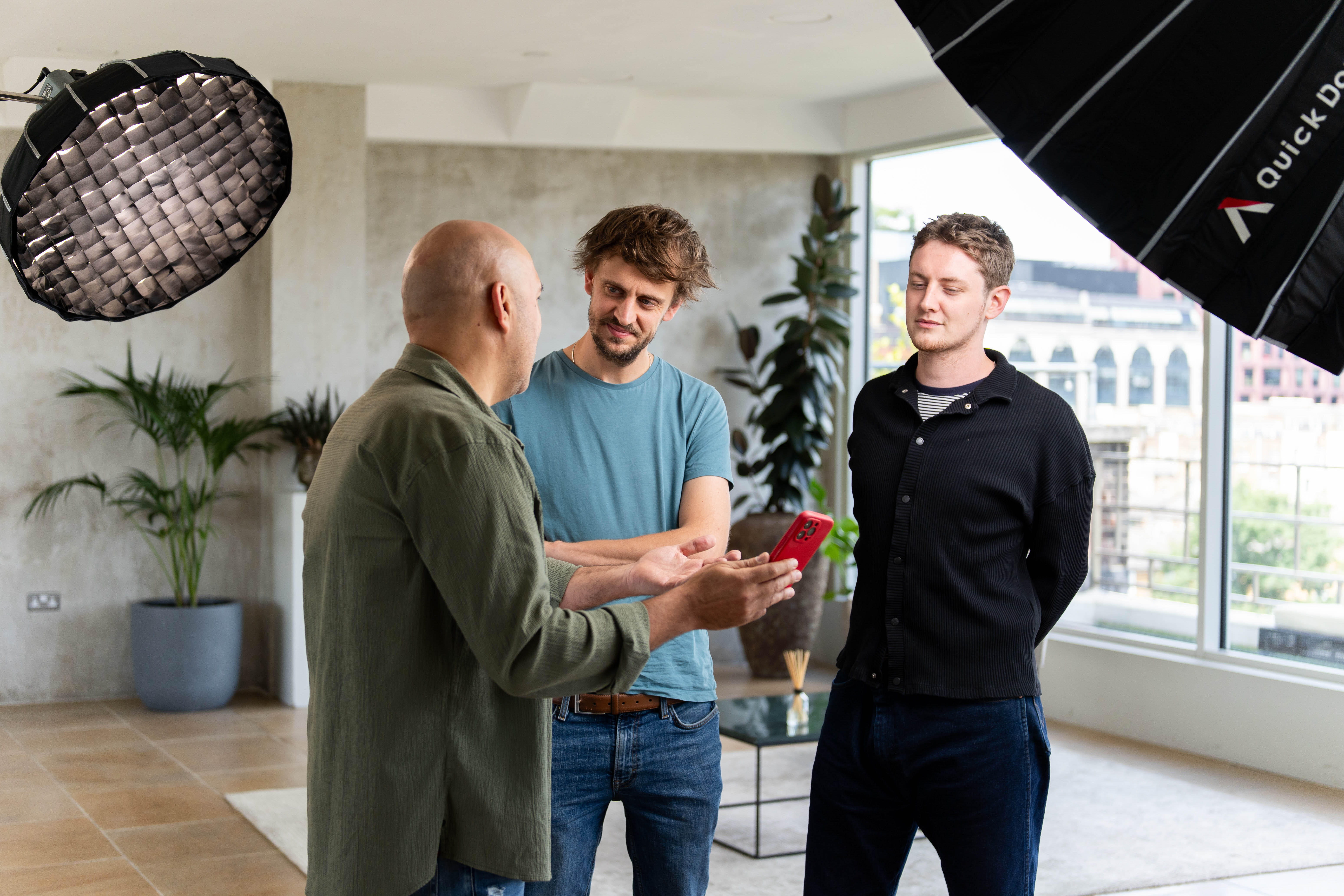 Three men standing and talking between takes on a video shoot set, with one holding a phone, surrounded by softbox lighting rigs and large windows overlooking a city skyline.