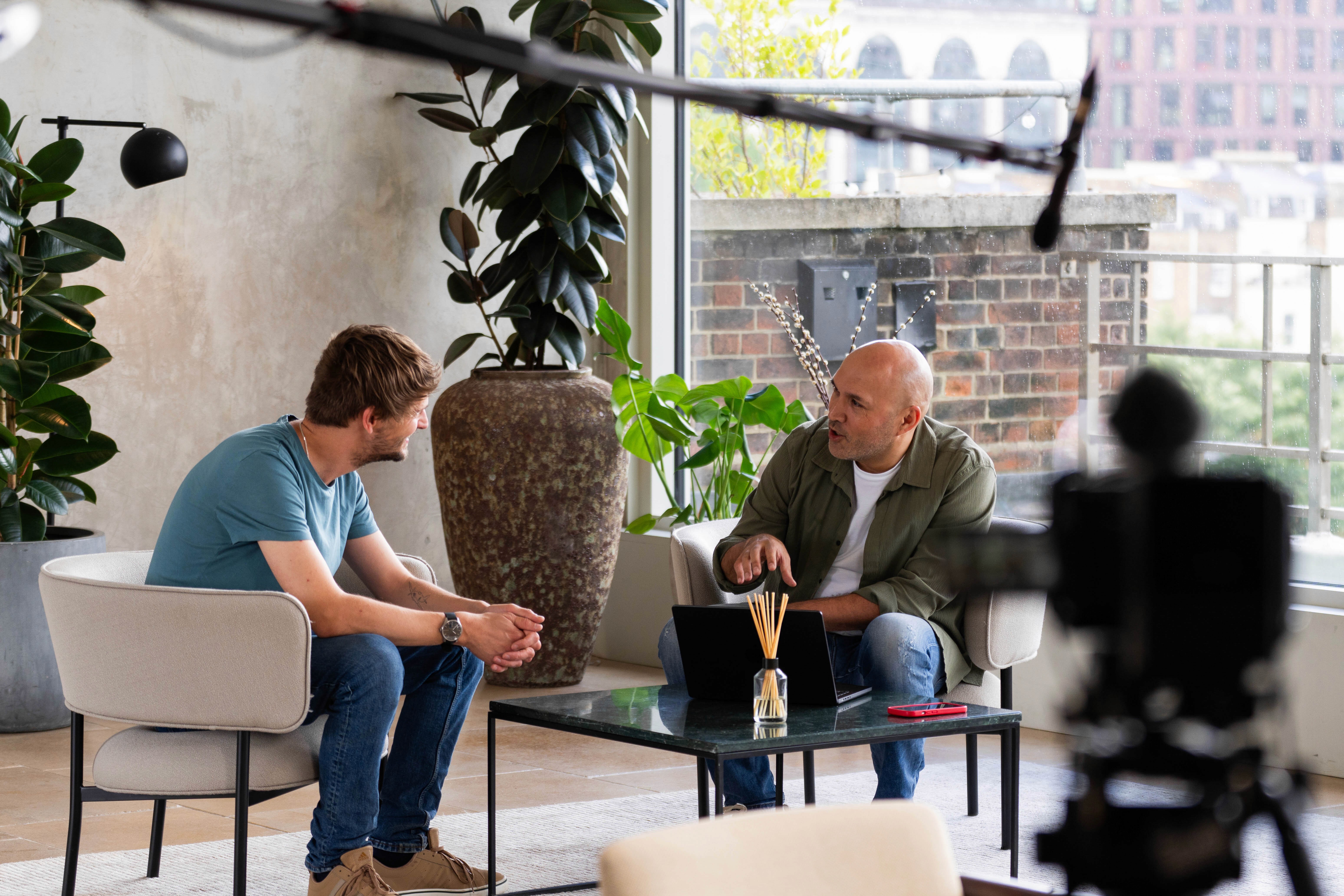 Two men seated across from each other in a bright studio space having a filmed conversation, with a laptop and reed diffuser on a glass coffee table between them, a camera and boom microphone visible in the foreground.