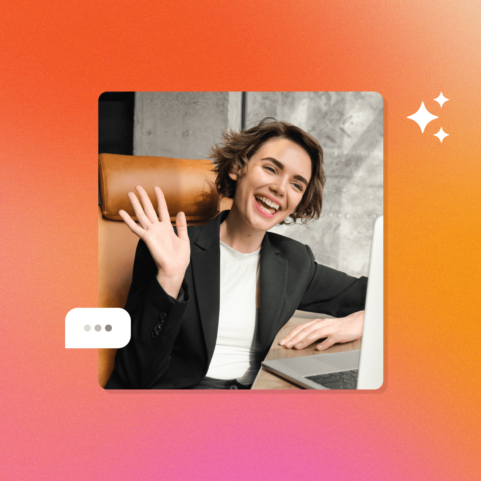 A smiling woman in a black blazer waves at a laptop screen while seated in a leather chair, with a white speech bubble showing an ellipsis to her lower left and a white sparkle icon to her upper right, all set against a warm orange-to-pink gradient background.