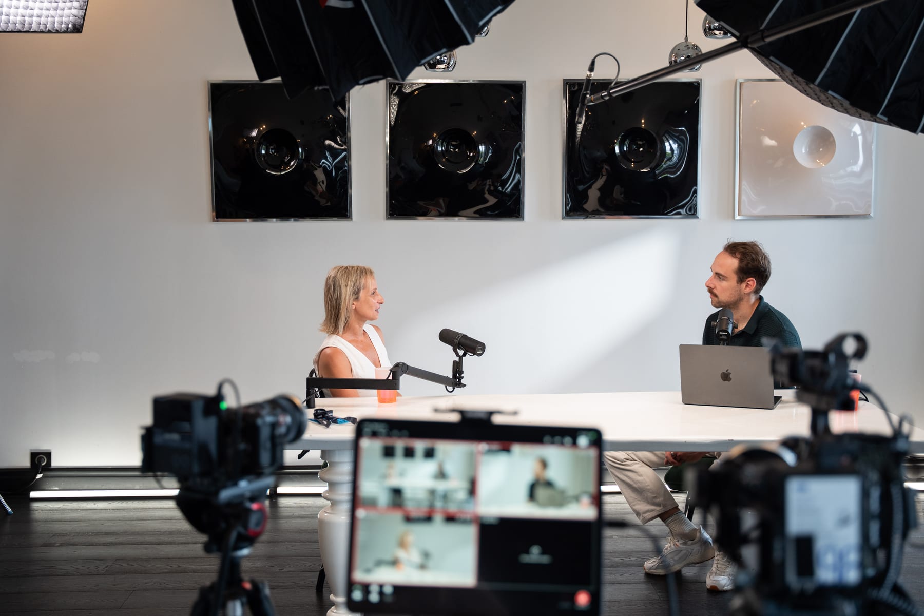 Angeliki Maragkou and host Tom Husson in conversation at the white table, seen past cameras and a multi-angle monitor on the Conveo podcast set at Cannes Lions 2026