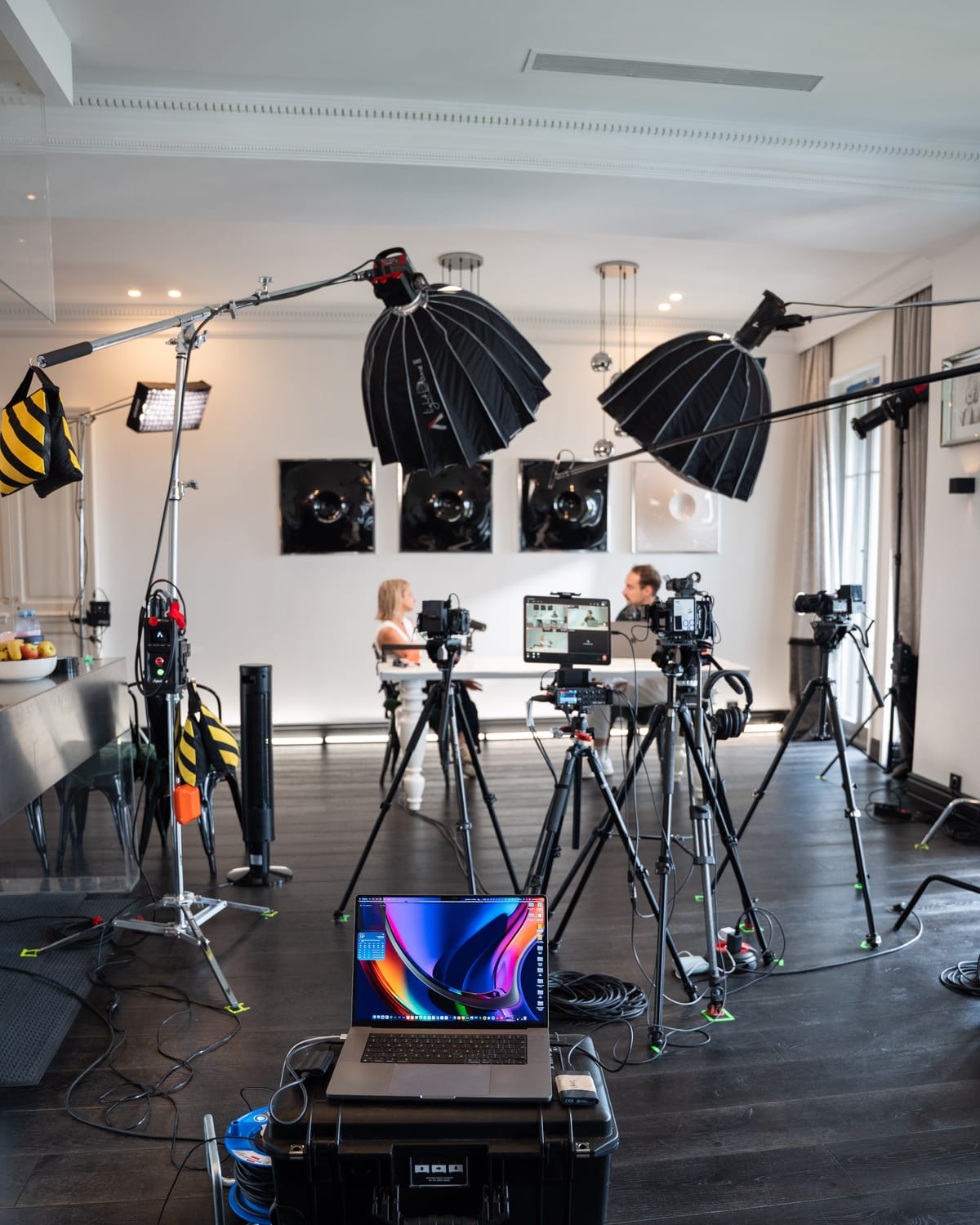 Wide behind-the-scenes view of cameras, lights, cables and a laptop on flight cases, with Maragkou and Husson recording at the table in the background at Cannes Lions 2026