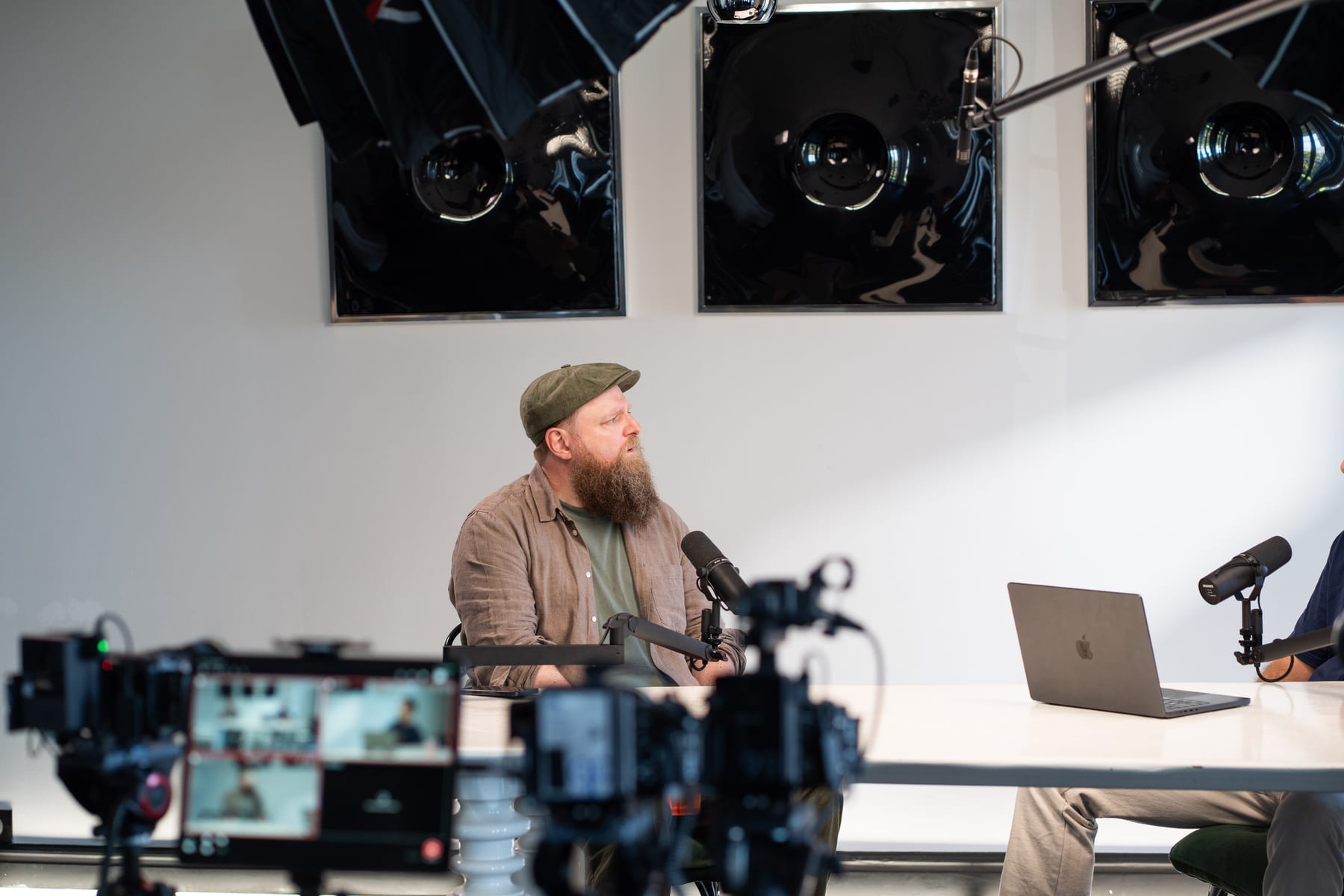 Wesley ter Haar in a green cap and beard looks across the table toward the host, seen past camera gear on the Conveo podcast set at Cannes Lions 2026