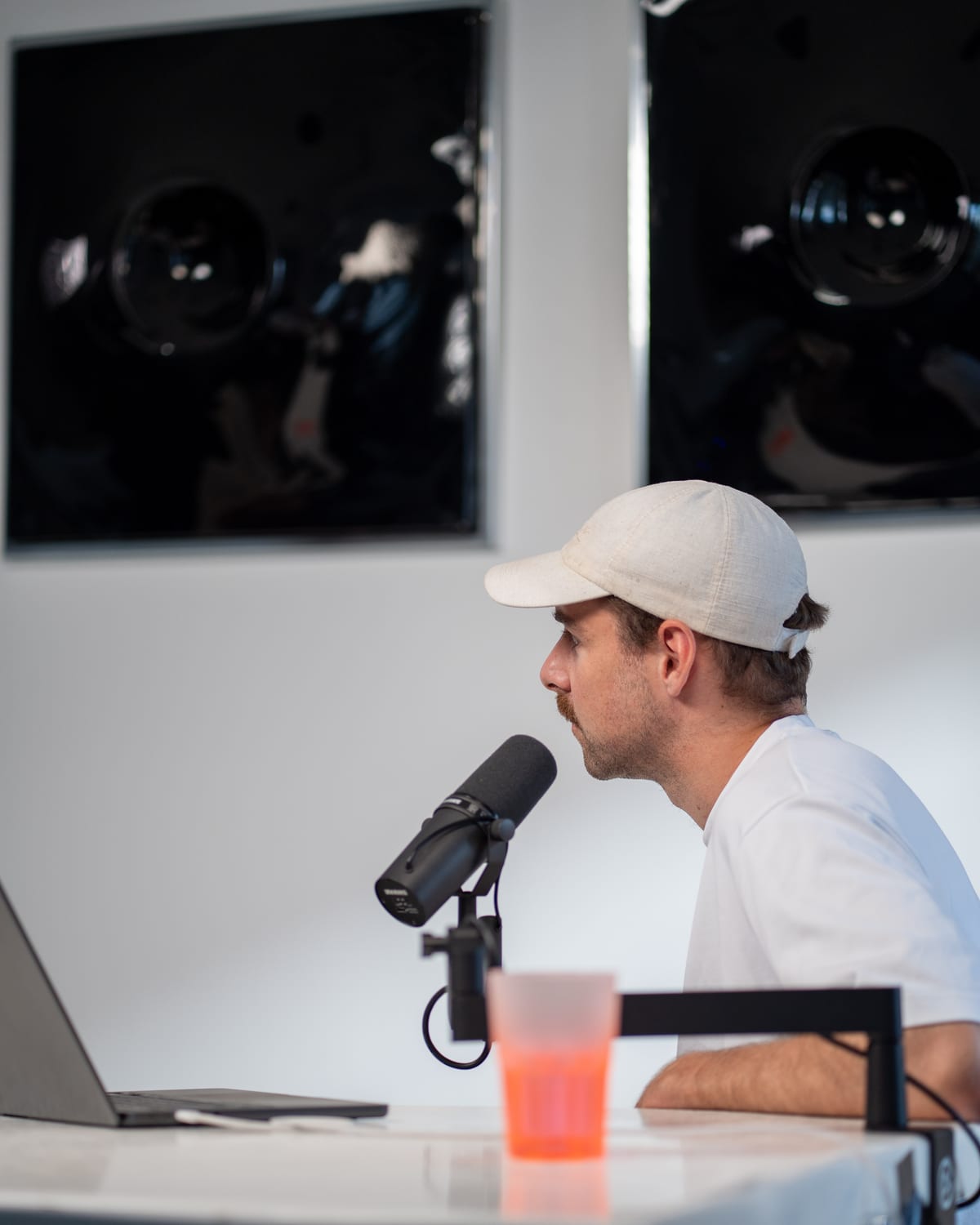 The host, in a white cap, gestures while talking with Brock Johnson at the microphone on the Conveo podcast set at Cannes Lions 2026.