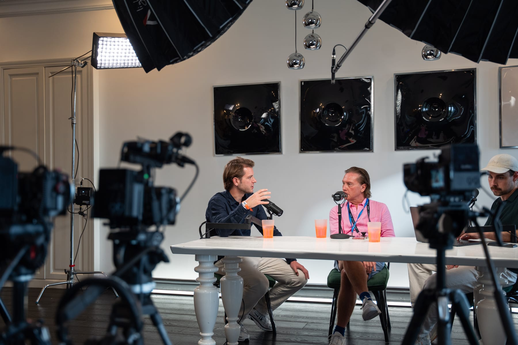 A host gestures toward Christopher Nurko across the white table on the Conveo podcast set, with video cameras and studio lights in the foreground at Cannes Lions 2026.