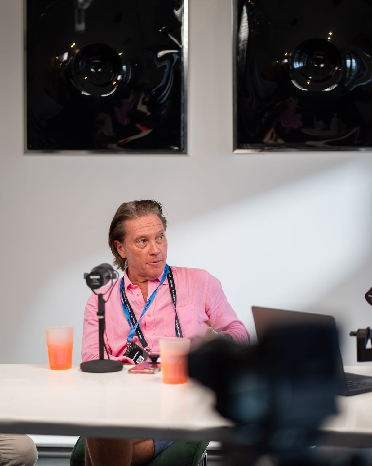 Christopher Nurko in a pink shirt and festival lanyard turns toward a host at the microphone on the Conveo podcast set at Cannes Lions 2026.