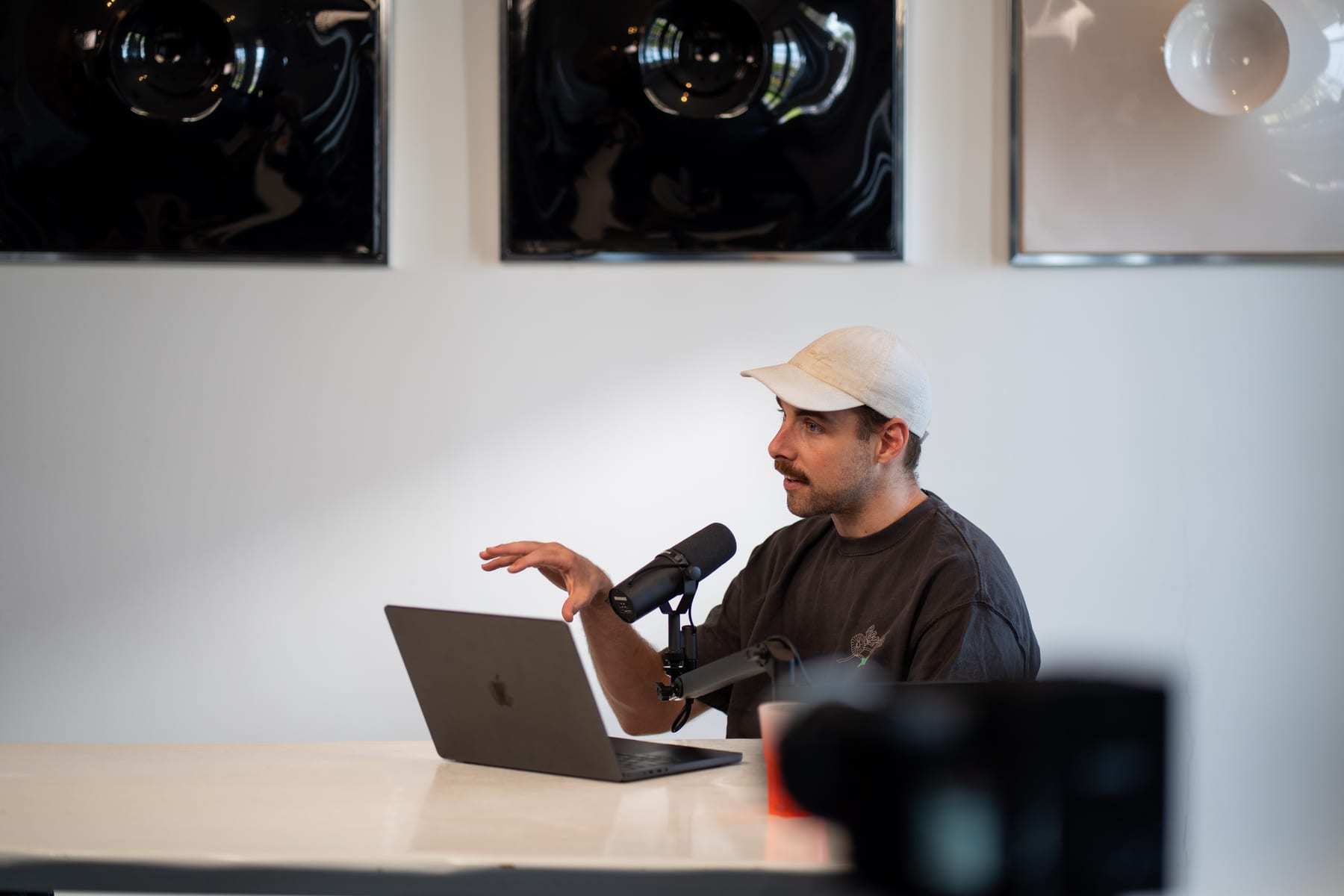 The host in a white cap gesturing as he speaks into a microphone behind his laptop on the Conveo podcast set at Cannes Lions 2026
