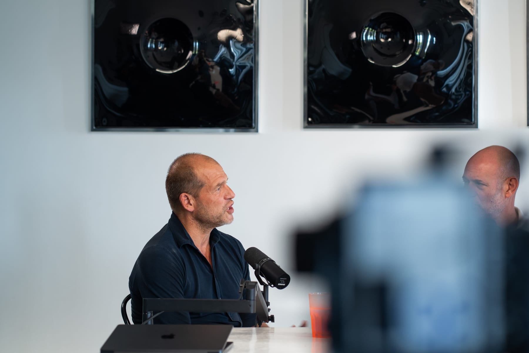 Kees Kruythoff in a dark shirt speaking into a microphone at the podcast table, with a blurred camera in the foreground, at Cannes Lions 2026
