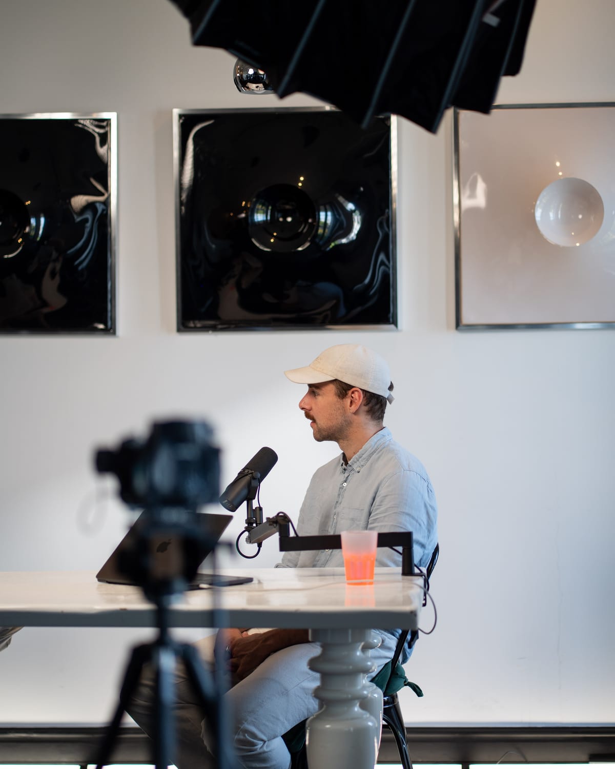 The Conveo host in a white cap and light blue shirt asks a question into his microphone, seen past a camera on a tripod at Cannes Lions 2026