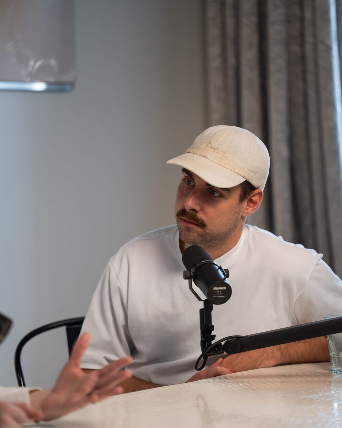 The host in a white cap and white T-shirt speaks at the microphone, gesturing across the table, at Cannes Lions 2026