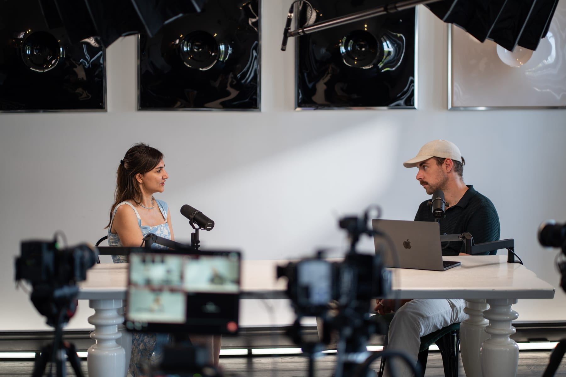 Özge Akcizmeci and the host face each other across the podcast table, framed by cameras and a monitor in the foreground at Cannes Lions 2026