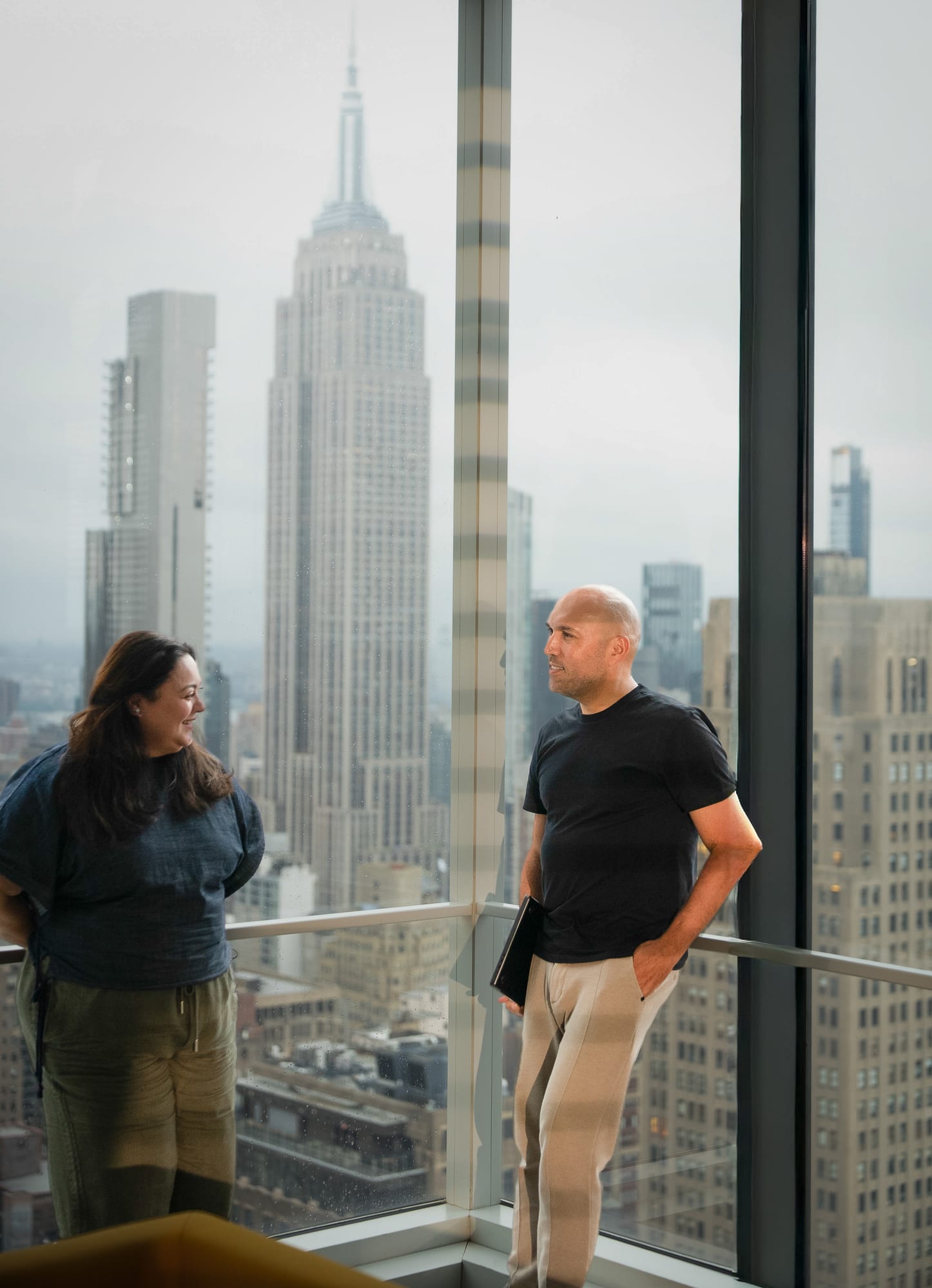 Carla Mahfuz and Rómulo Rejón talking by a floor-to-ceiling window high above Manhattan, the Empire State Building behind them.