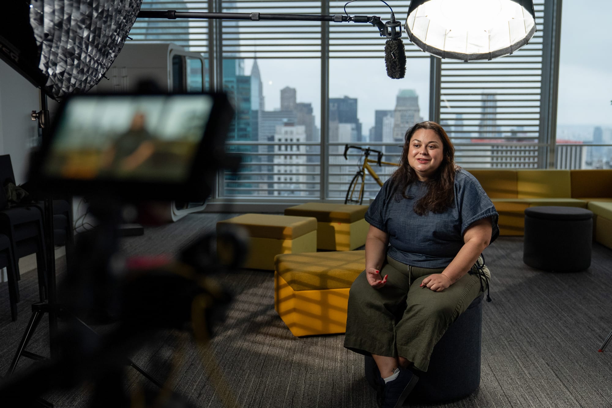 Carla Mahfuz seated on a stool mid-interview in a bright office with the Manhattan skyline behind her, seen past a camera monitor, boom microphone and softbox light in the foreground.