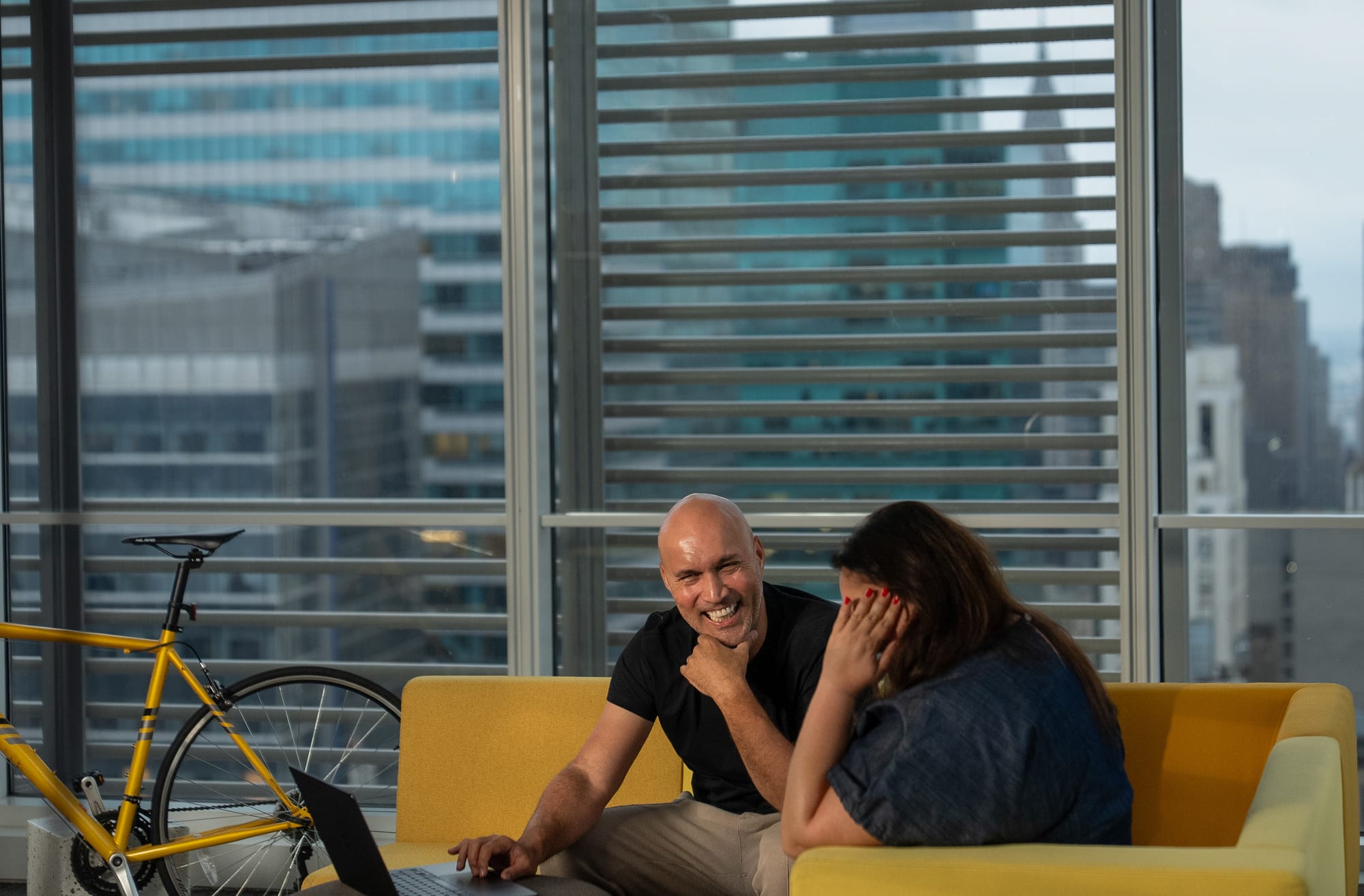 Carla Mahfuz and Rómulo Rejón laughing on a yellow sofa in front of a window onto Midtown Manhattan, a laptop open between them and a yellow bicycle to the side.