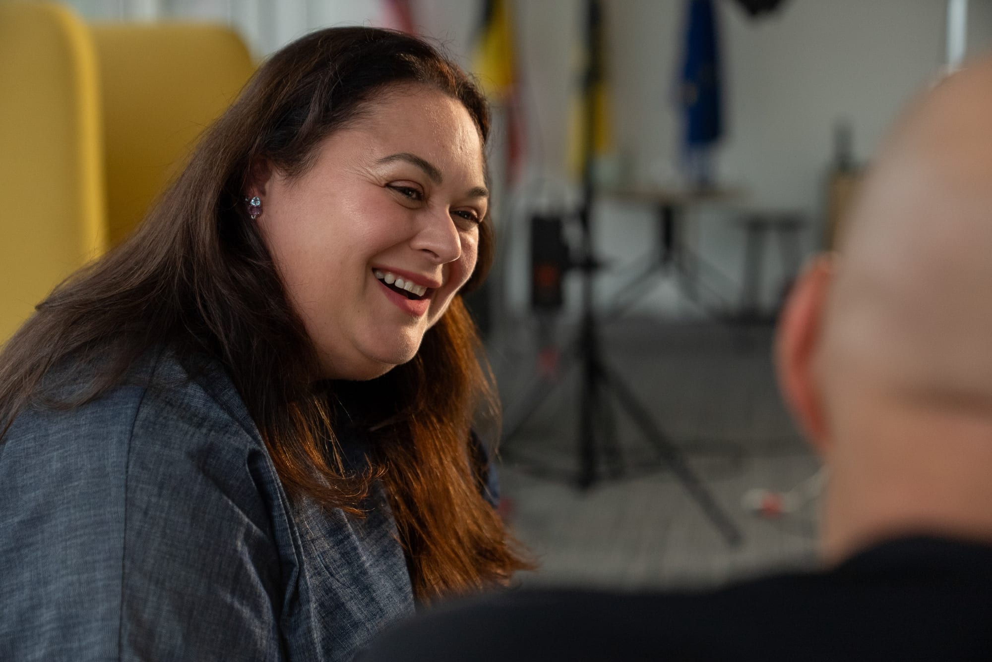 Close-up of Carla Mahfuz laughing mid-conversation, long dark hair over a gray top, with a yellow sofa and a row of flags out of focus behind her.
