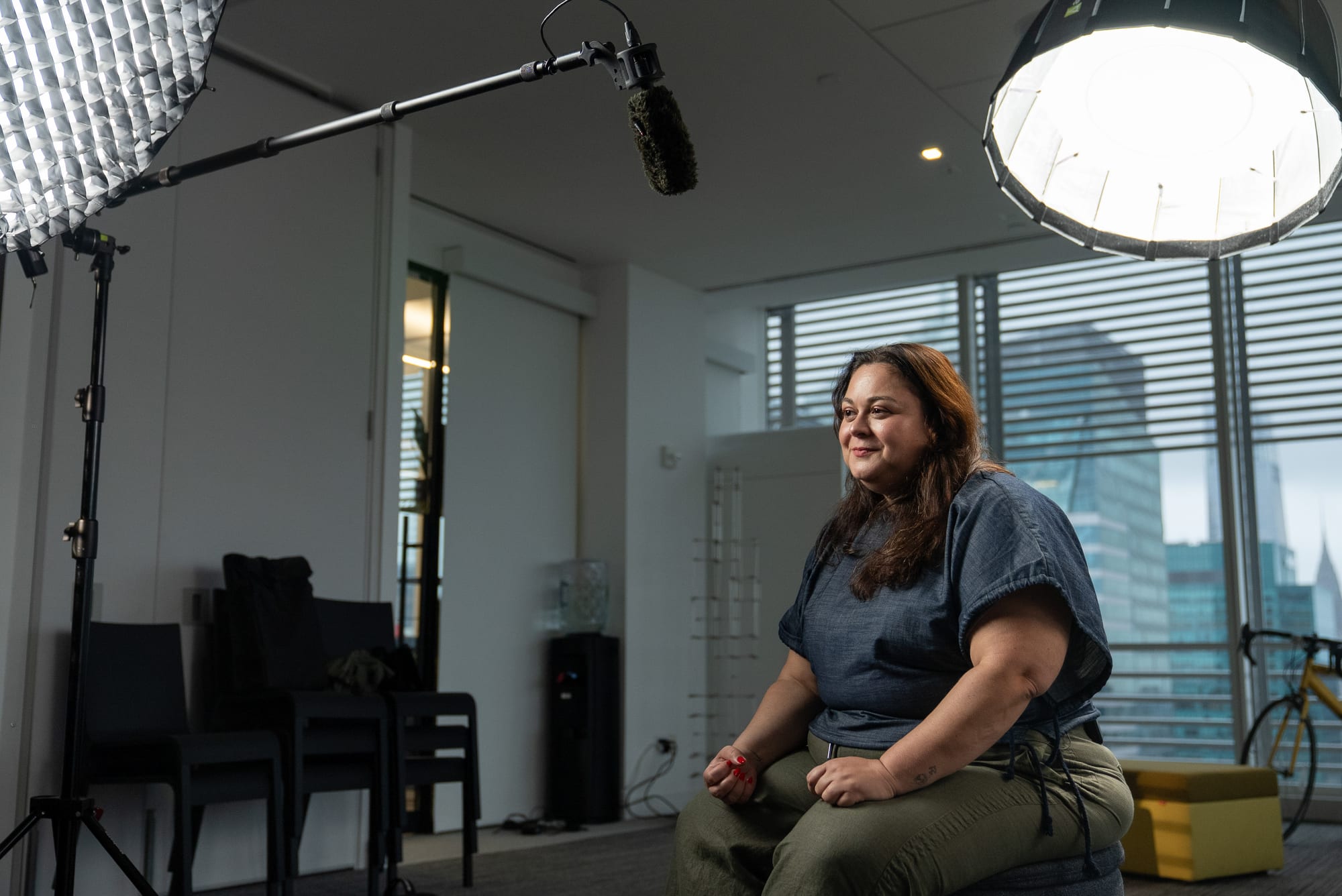 Carla Mahfuz seated for her interview under a softbox light and boom microphone, smiling off camera, with Midtown towers through the window behind her.