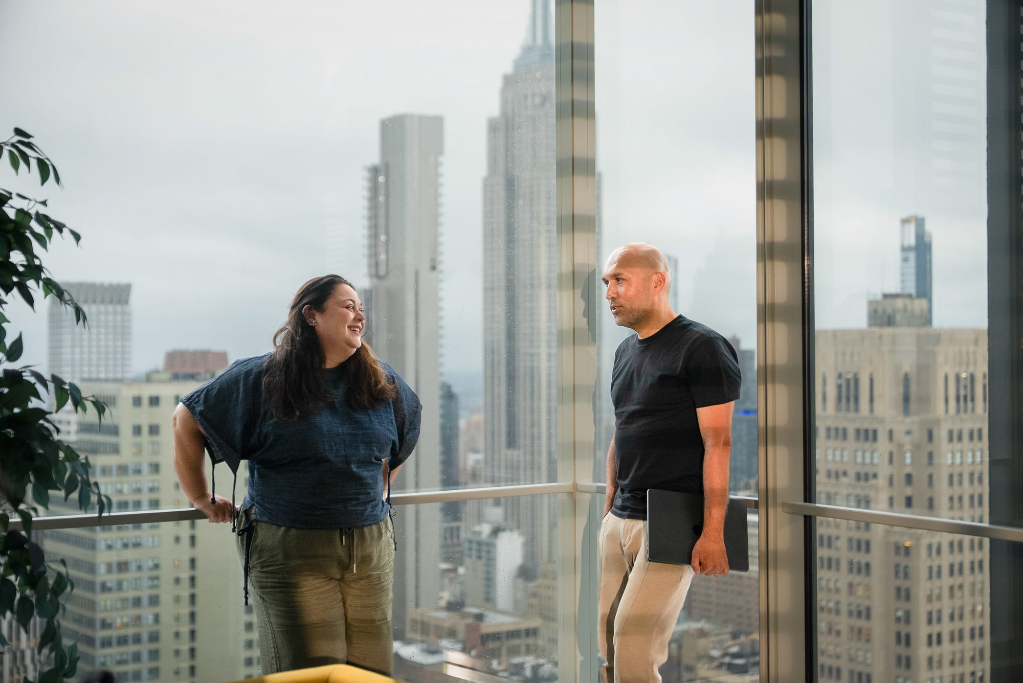 Carla Mahfuz and Rómulo Rejón standing at a corner window high above Manhattan, the Empire State Building between them in the skyline.