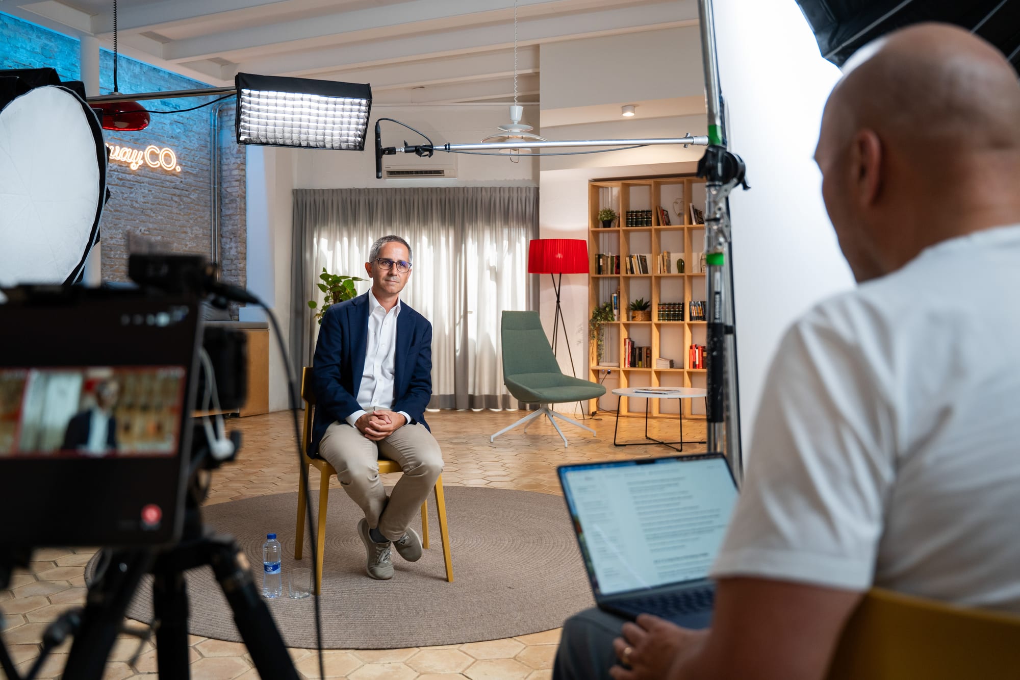 Wide view of the interview set: Pablo Pérez seated under studio lights, a camera rig and monitor in front of him, a colleague at a laptop in the foreground.
