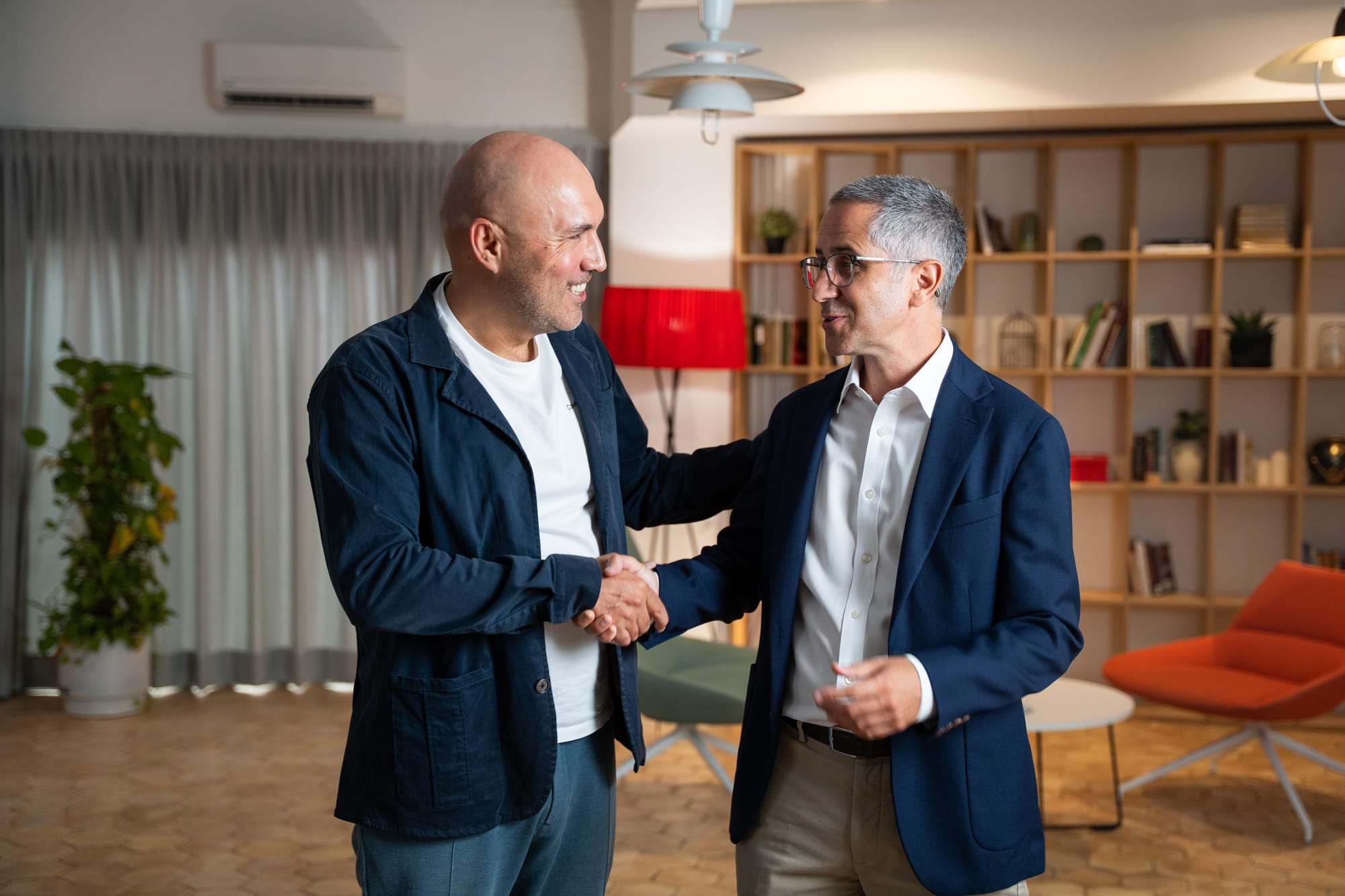 Pablo Pérez and a Conveo colleague shaking hands and smiling on the interview set, bookshelves and a red lamp behind them.