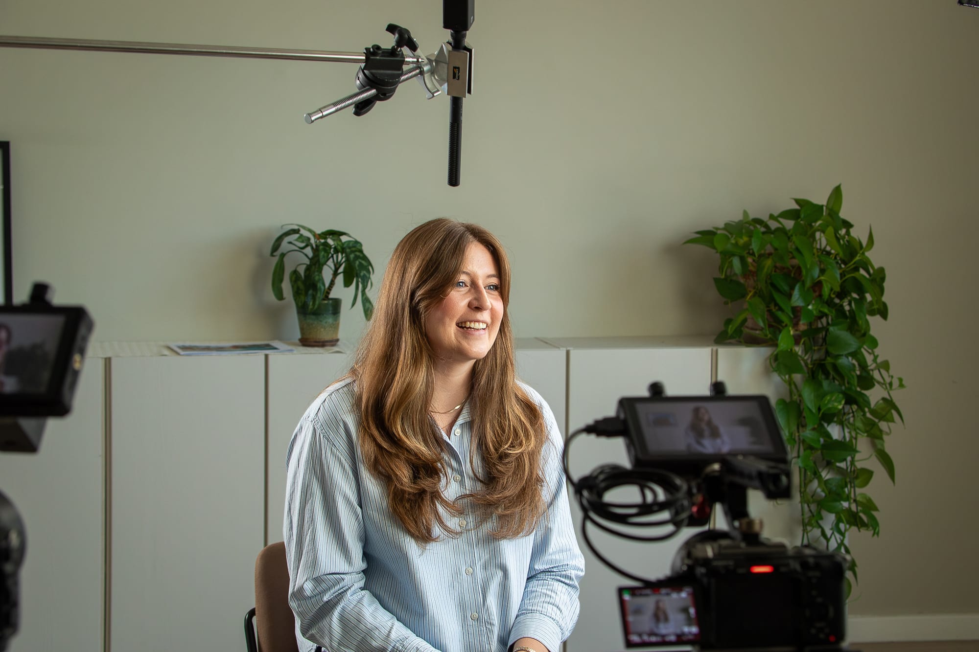 Chloe Cook, Market Insights Manager at HeliosX, smiling in a light blue striped shirt during a video shoot, with a boom microphone overhead, a camera monitor in the foreground and potted plants behind her.