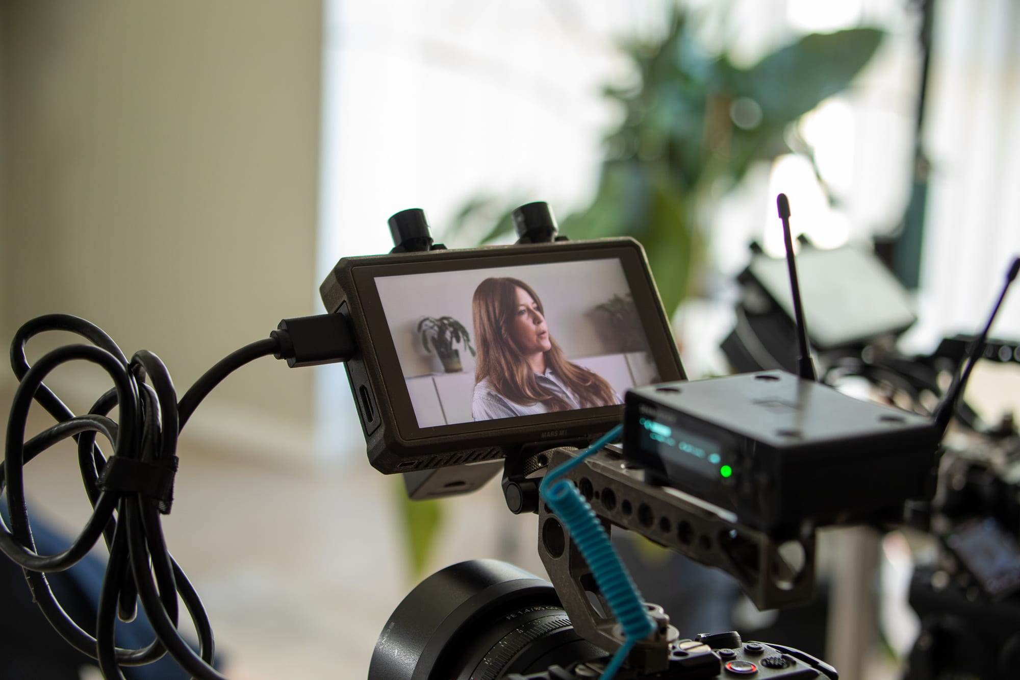 Close-up of a camera monitor showing Chloe Cook mid-interview, with the camera rig, coiled cables and a wireless transmitter in focus and the room blurred behind.