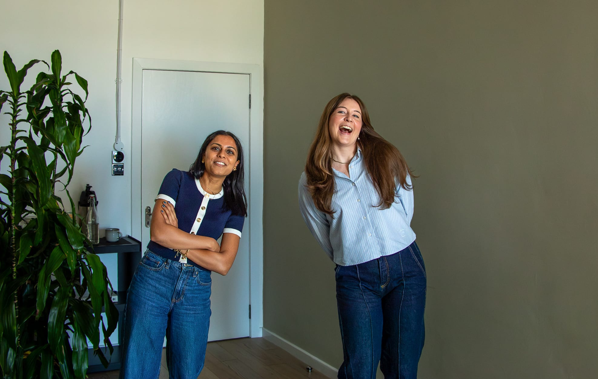 Chloe Cook and a colleague laughing together in a bright room with a tall plant, both in jeans, between takes on the video shoot.