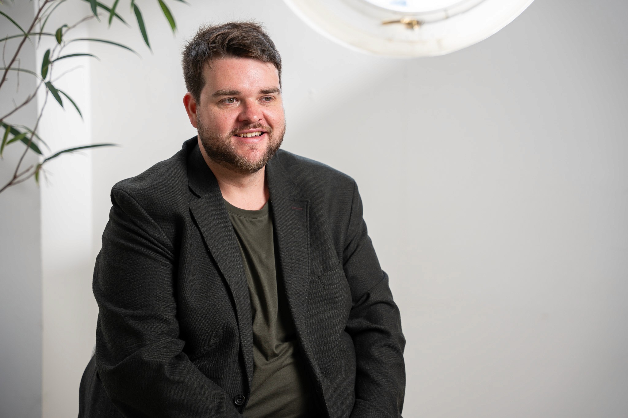 Portrait of a man in a dark blazer and olive t-shirt smiling in a bright studio space, with a potted plant and circular porthole window in the background.
